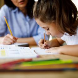 Young girl writing in workbook while adult watches